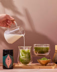 A hand pours milk into a glass of iced Kagoshima matcha. Nearby are a jar of Organic Ceremonial Matcha 700 powder, a bowl of tea, a bamboo whisk, a scoop with green powder, and a fine mesh strainer on a tray.