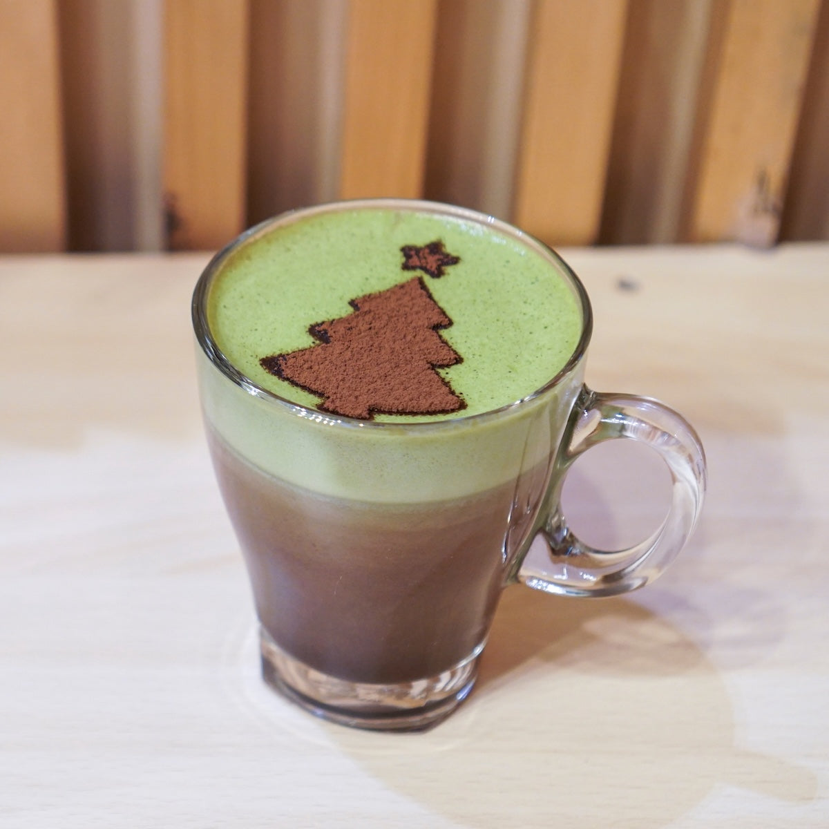 A glass mug of Mint Matcha Cocoa, with green and brown layers and cocoa powder dusted in a Christmas tree with a star, sits on a light wood table against a wooden background.