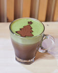 A glass mug of Mint Matcha Cocoa, with green and brown layers and cocoa powder dusted in a Christmas tree with a star, sits on a light wood table against a wooden background.