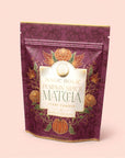 A purple resealable pouch labeled “Pumpkin Spice Matcha,” adorned with gold accents, orange flowers, and a pumpkin illustration on the front, set against a light pink background—this elegant packaging highlights autumn botanicals and features the rich flavor of ceremonial matcha expertly blended with warming pumpkin spice.