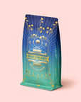 A colorful bag labeled "Mercury Mint Nighttime: Vanilla Mint Lavender Jasmine Tea Refill Pouch" features intricate gold designs, yellow flowers, and celestial motifs on a blue-to-green gradient background, set against a soft pink surface—perfect for anyone seeking soothing nighttime digestion support with this herbal tea blend.