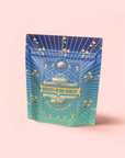A colorful resealable pouch labeled "Mercury Mint Nighttime: Vanilla Mint Lavender Jasmine Tea - Caffeine Free!" with gold and turquoise astrological motifs and sunbursts offers a soothing herbal blend, set against a pastel pink background.
