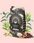 A patterned bag labeled "Metabolism Balance: Breakfast with Benefits" rests among ginger, tea leaves, and leafy greens on a pink background, evoking a natural, health-focused metabolism tea inspired by adaptogen ingredients.