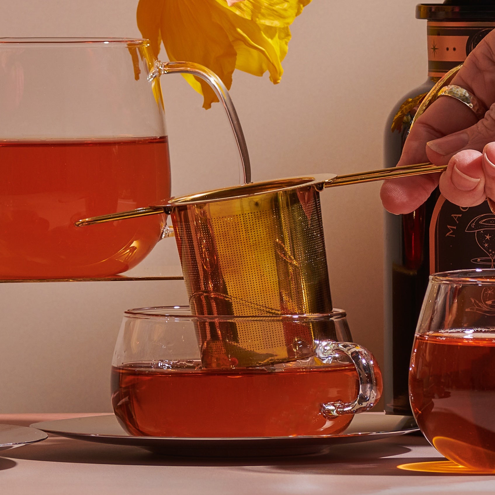 A hand is seen pouring tea through a Midas Touch: Golden-Hued Tea Strainer by PMB Mesh Screen Company into a glass teacup. A teapot filled with amber liquid is also visible. Another teacup is partially seen in the foreground. A daffodil in a glass container and a dark bottle are in the background.