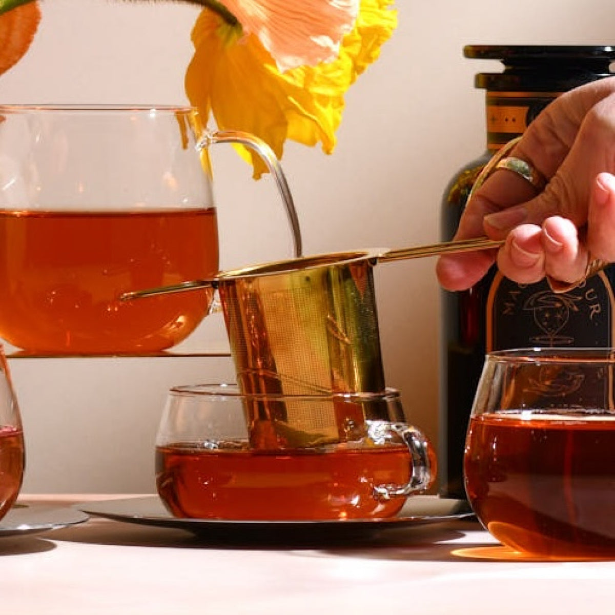 A hand holding an artisan-crafted gold tea strainer from the Magic Hour Starter Kit by Magic Hour is pouring tea into a glass cup on a saucer. Another glass teacup and a small glass teapot containing amber tea are on the surface. Bright orange flowers and a dark amber bottle in the background exude the elegance of a traditional tea ceremony.