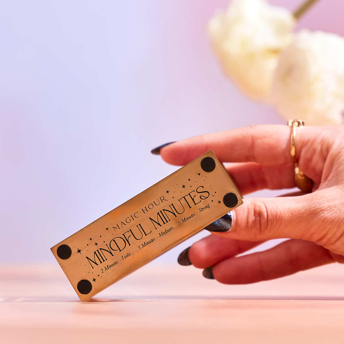 A hand with dark nail polish holds the Mindful Minutes Tea Timer box, labeled "MAGIC HOUR MINDFUL MINUTES," against a soft pink background with blurred white flowers, highlighting its elegant design for precision tea steeping.