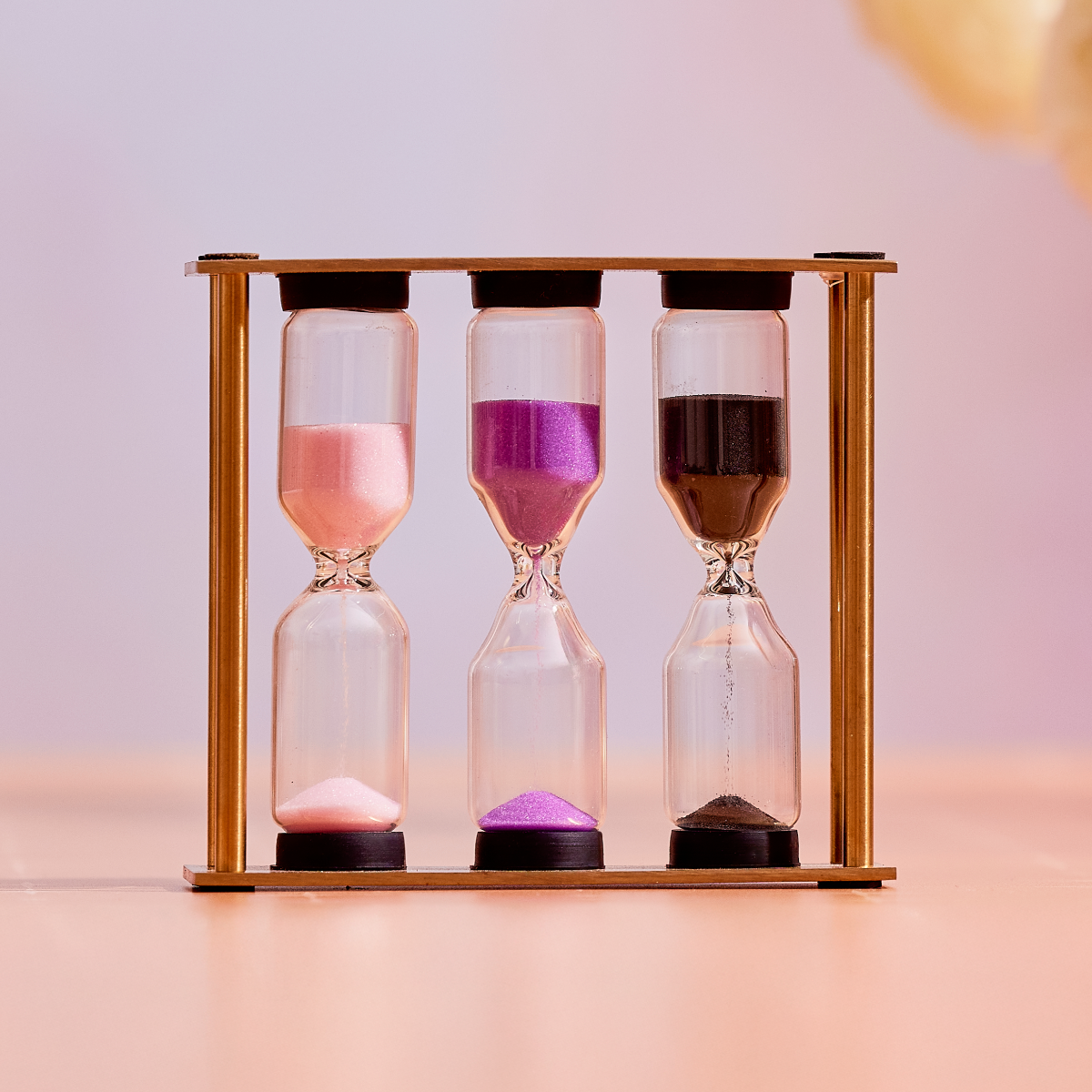 The Mindful Minutes Tea Timer features a wooden frame holding three hourglasses with pink, purple, and black sand, each partially emptied for precise tea steeping, set against a soft pink background.