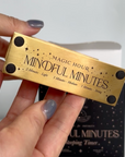 A person with gray nail polish holds a rectangular brass item labeled "Mindful Minutes Tea Timer." The item is marked with intervals: 2 Minutes - Light, 3 Minutes - Medium, 5 Minutes - Strong. The background includes a partially open box with similar branding from Alibaba.