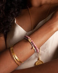 A person with curly hair wears a white spaghetti strap top, displaying gold bangles, the Rose Quartz Mini Hamsa Bracelet, and two beaded stretch bracelets in pink and purple. The arms are crossed, highlighting the jewelry against the background of light-colored clothing.