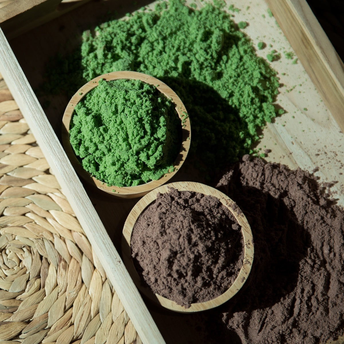 Two wooden bowls sit on a tray: one holds vibrant Mint Matcha Cocoa and the other, rich chocolate mint powder. Some powder is scattered nearby, and a woven mat is partially visible beside the tray.