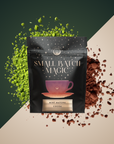 A black "Small Batch Magic" cocoa package labeled "Mint Matcha Cocoa," with a purple teacup design, sits atop scattered green matcha and brown cocoa powder on a two-tone background.
