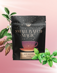 A bag labeled "Small Batch Magic" with "Mint Matcha Cocoa" sits centered on a pink gradient background, surrounded by illustrated cocoa pods, mint leaves, and blooming branches—capturing the spirit of Mint Matcha Cocoa.