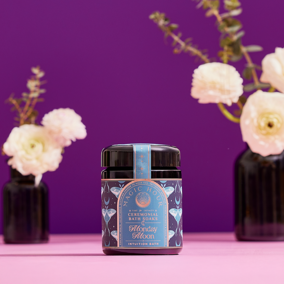 A jar of Ceremonial Bath Salt Soaks - Violet Glass Jar rests on a pink surface with white flowers in black vases and a purple background, its blue celestial-themed label inviting a cosmic touch to your self-care ritual.