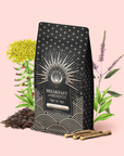 A black coffee bag labeled "Mood Balance: Breakfast with Benefits" stands among herbs and roots—featuring yellow, purple, and green plants—to evoke the nourishing qualities of an Ashwagandha or Lion's Mane tea blend, set against a pale pink background.