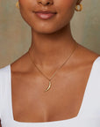 A person wearing a white sleeveless top, gold hoop earrings, and the Midnight Sky Pendant Moon Necklace featuring a celestial realm-inspired hammered 18kt crescent moon. The background is a plain, light, and slightly textured wall.