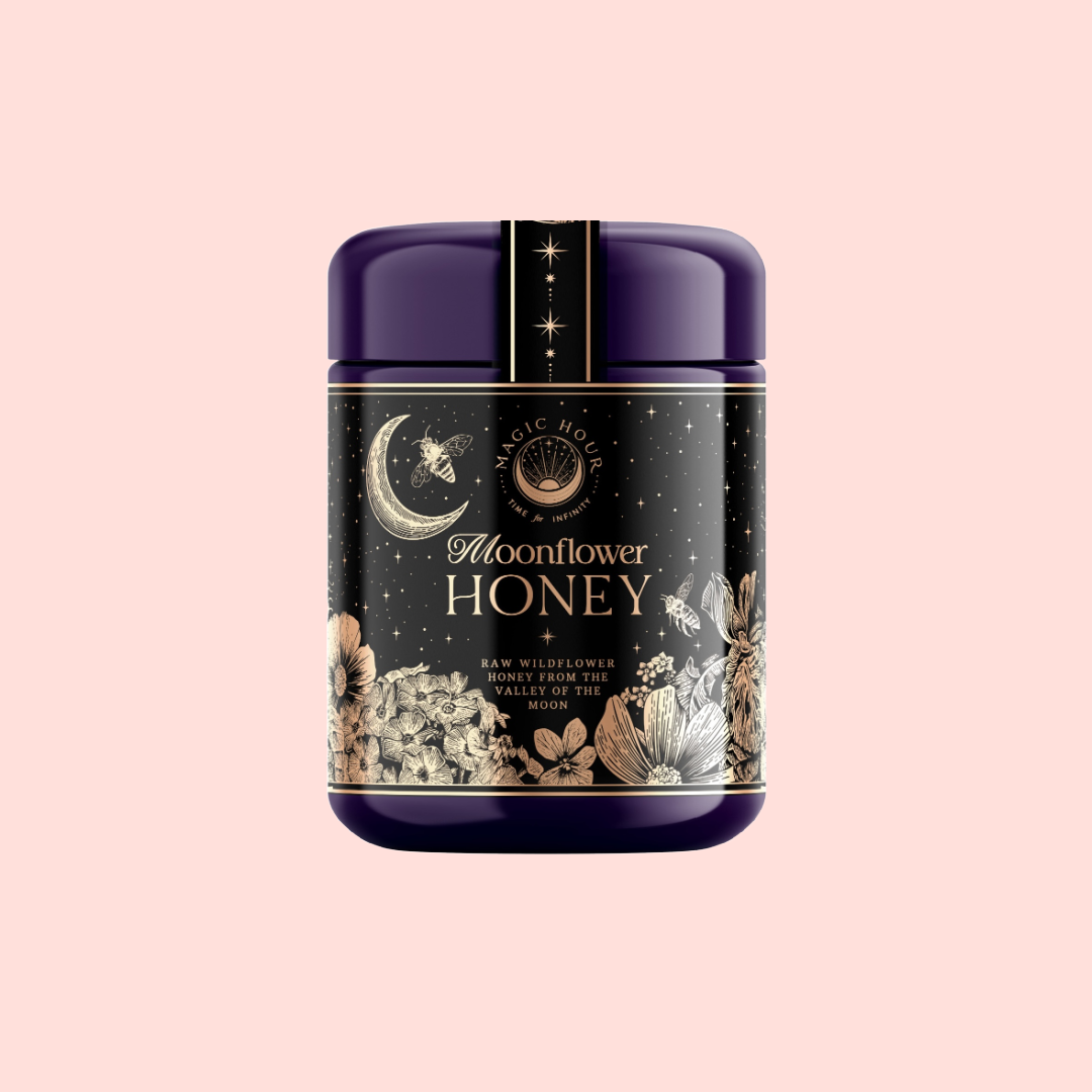 A jar of Moonflower Honey from Ojai Valley, adorned with a dark purple lid, showcases a celestial-themed label that features a crescent moon and stars. The label is set against a pale pink background and includes detailed floral illustrations that capture the essence of organic honey.