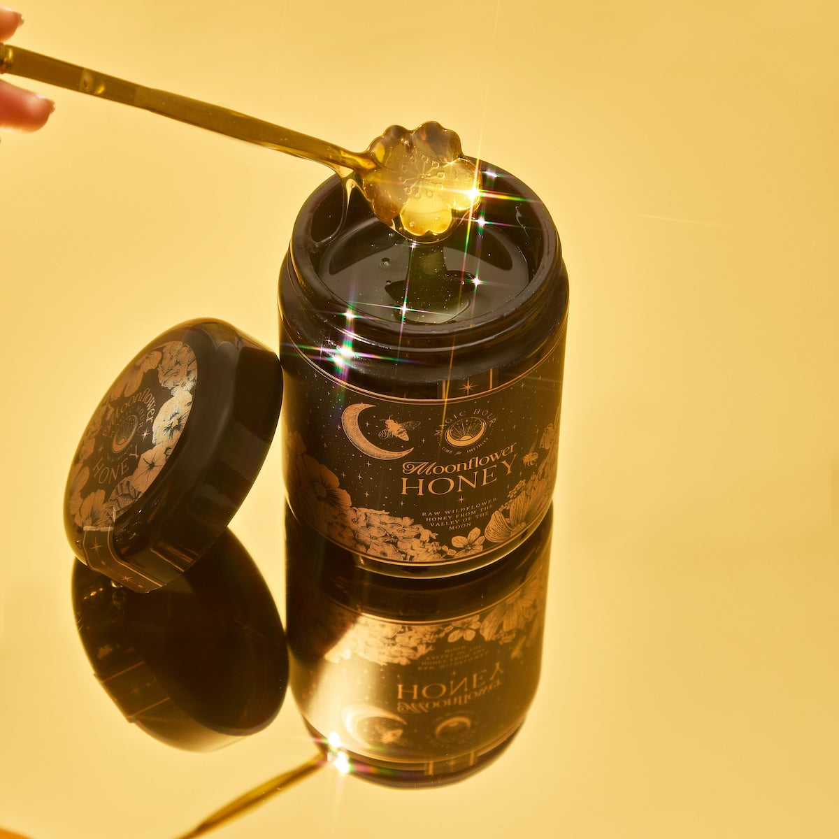 A jar of Moonflower Honey with a golden spoon lifting the antioxidant-rich honey. The celestial-designed jar and lid enhance its golden tones against a warm, bright yellow background.