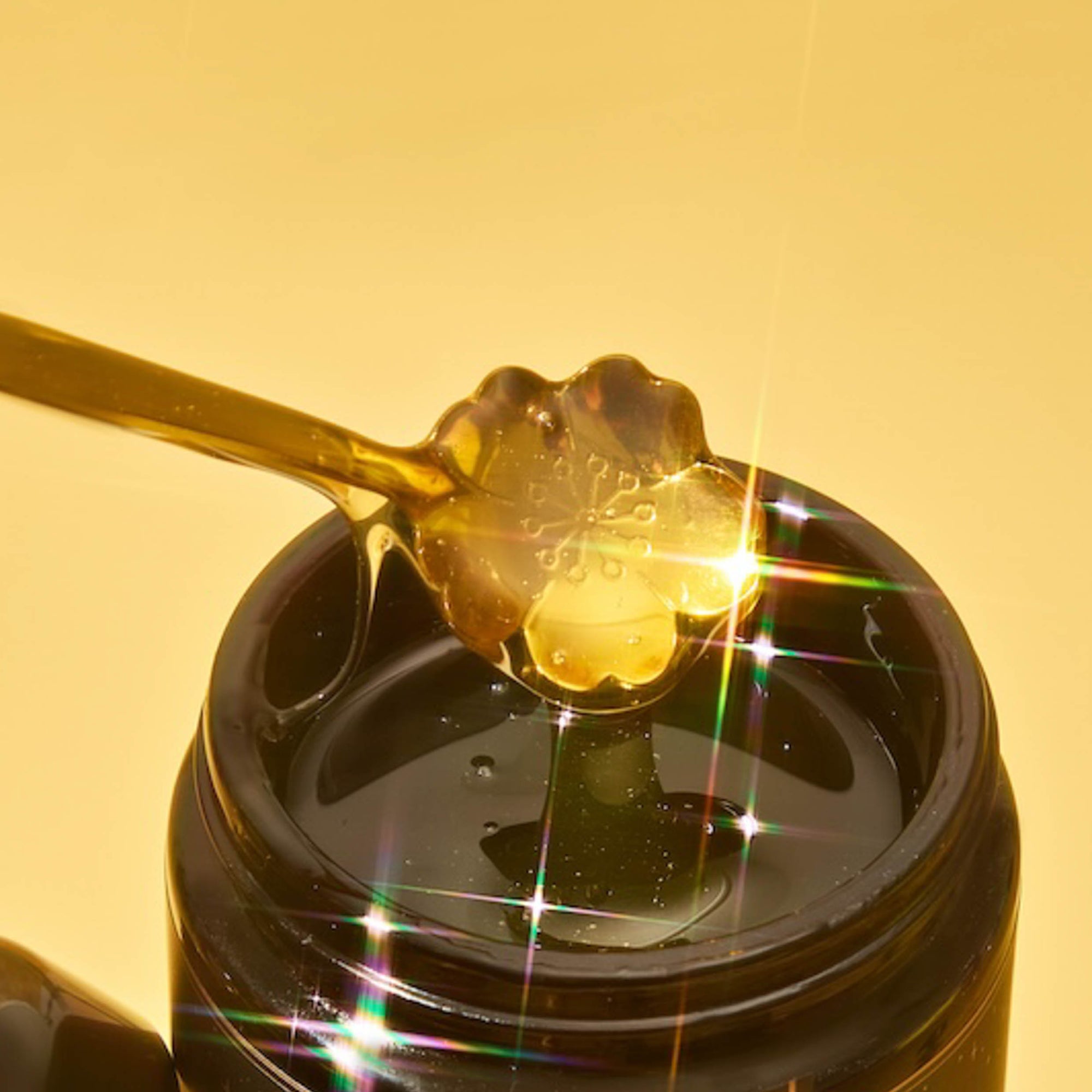 A Gold Floral Teaspoon drizzles golden, flower-shaped honey into an open jar, as bright light sparkles off the honey like stars against a yellow background.