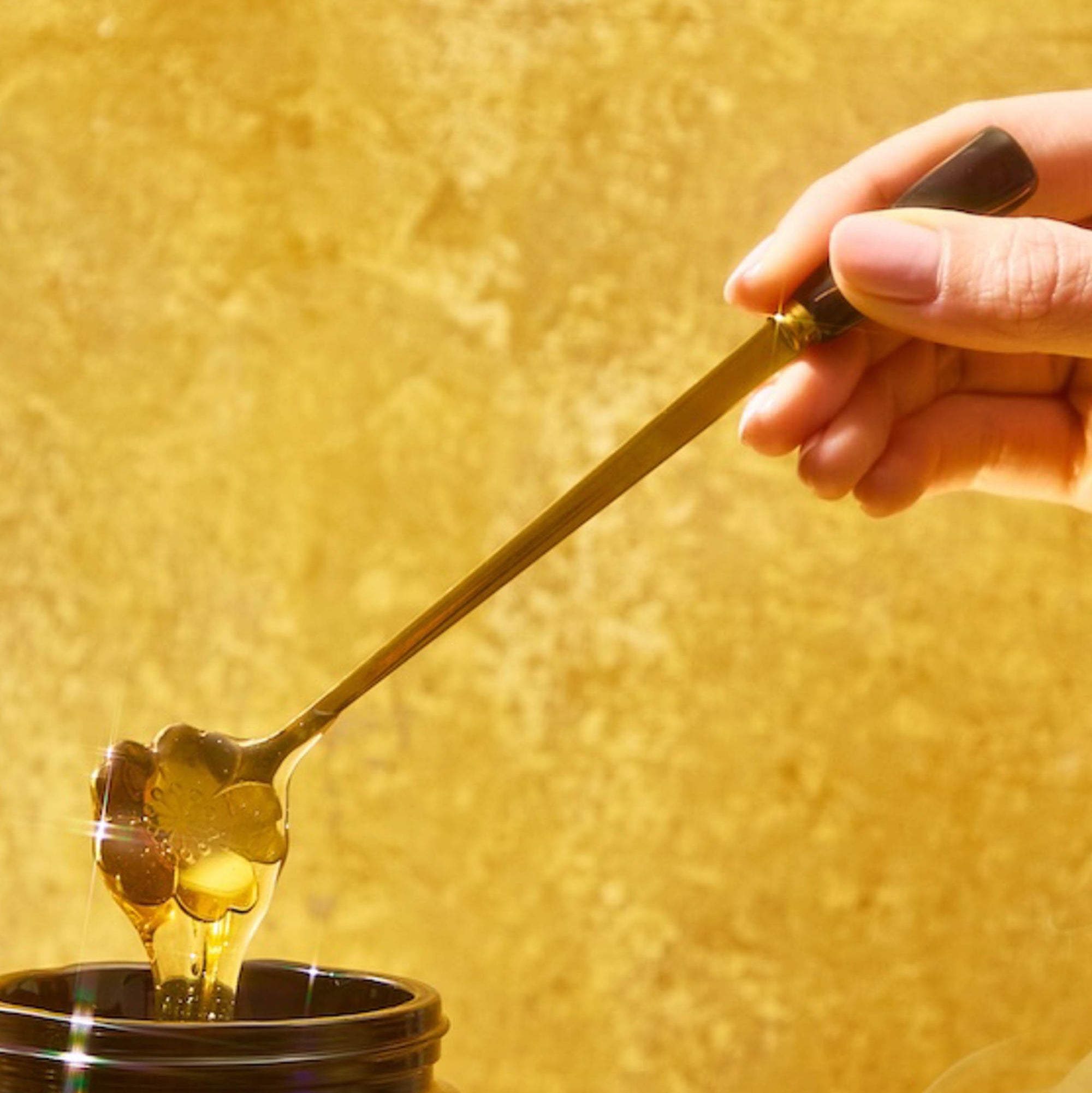 A hand holds a Gold Floral Teaspoon, lifting thick honey from a dark jar against a warm yellow background.