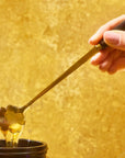 A hand holds a Gold Floral Teaspoon, lifting thick honey from a dark jar against a warm yellow background.
