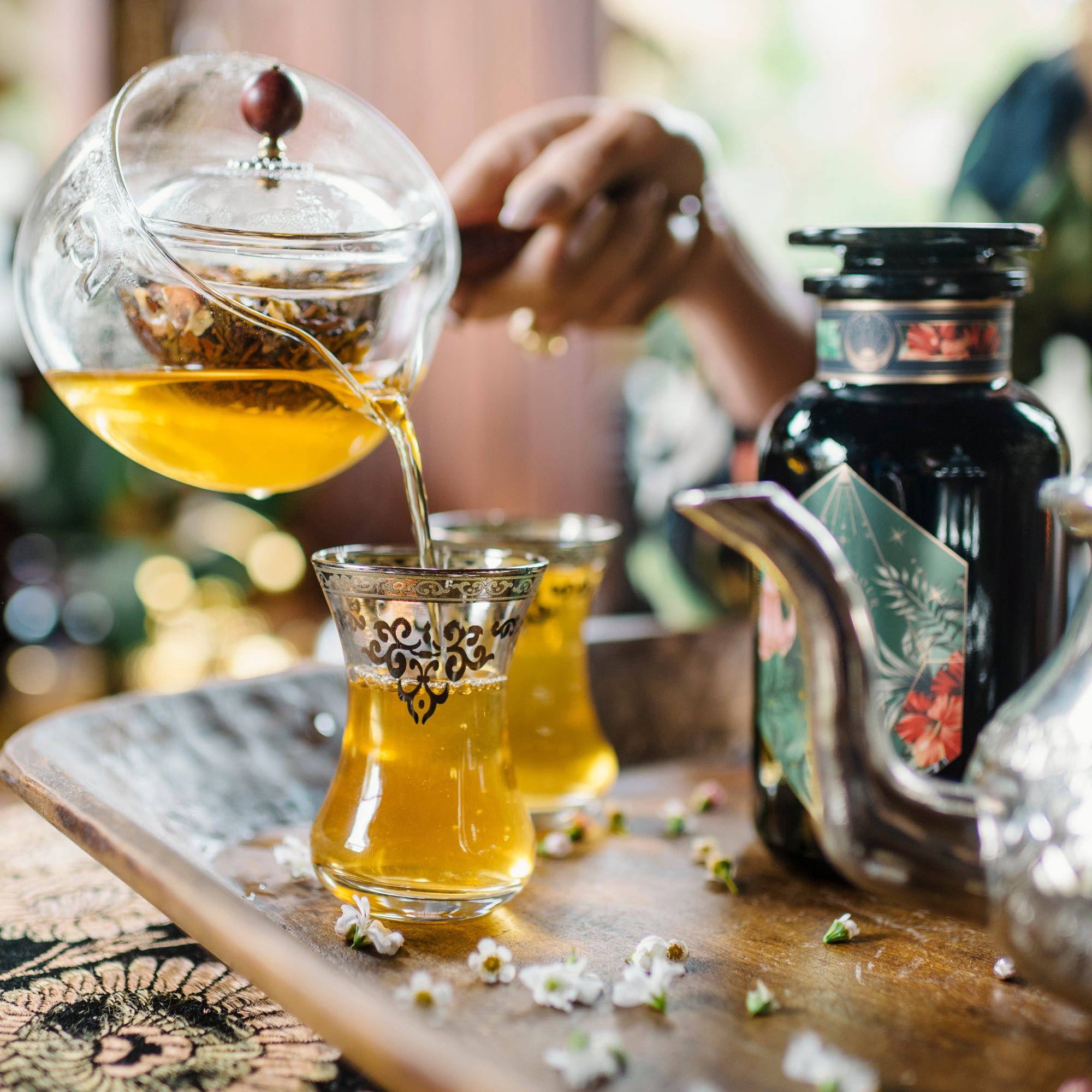 A hand pours Moonlight in Marrakesh: Spiced Mint Green Tea from a glass teapot into ornate glasses on a wooden tray, with flowers and a decorative black bottle nearby.