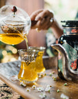A hand pours Moonlight in Marrakesh: Spiced Mint Green Tea from a glass teapot into ornate glasses on a wooden tray, with flowers and a decorative black bottle nearby.