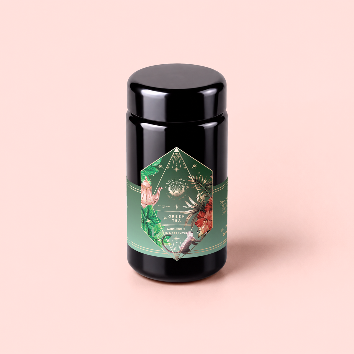 A canister of Moonlight in Marrakesh: Spiced Mint Green Tea, adorned with a green floral label and botanical illustrations, stands upright against a soft pink background—perfect for storing your favorite spiced herbal tea or Moroccan mint tea blends.
