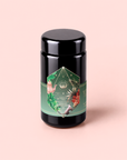 A canister of Moonlight in Marrakesh: Spiced Mint Green Tea, adorned with a green floral label and botanical illustrations, stands upright against a soft pink background—perfect for storing your favorite spiced herbal tea or Moroccan mint tea blends.