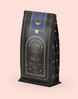 A standing dark bag with gold and blue accents displays the "Magic Hour" label, featuring an hourglass illustration. Star-like details hint at Moringa Moon Herbal Tea's digestive benefits. It rests on a light pink background.