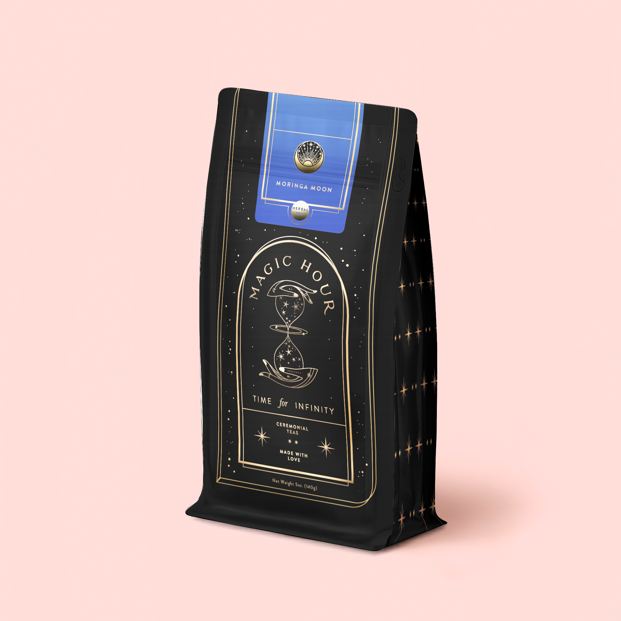 A black and gold coffee bag labeled “Magic Hour Time for Infinity,” adorned with an hourglass illustration, blue seal, and gold star patterns, rests against a pale pink background—capturing the enchanting appeal of the Moringa Moon Herbal Tea Refill Pouch.
