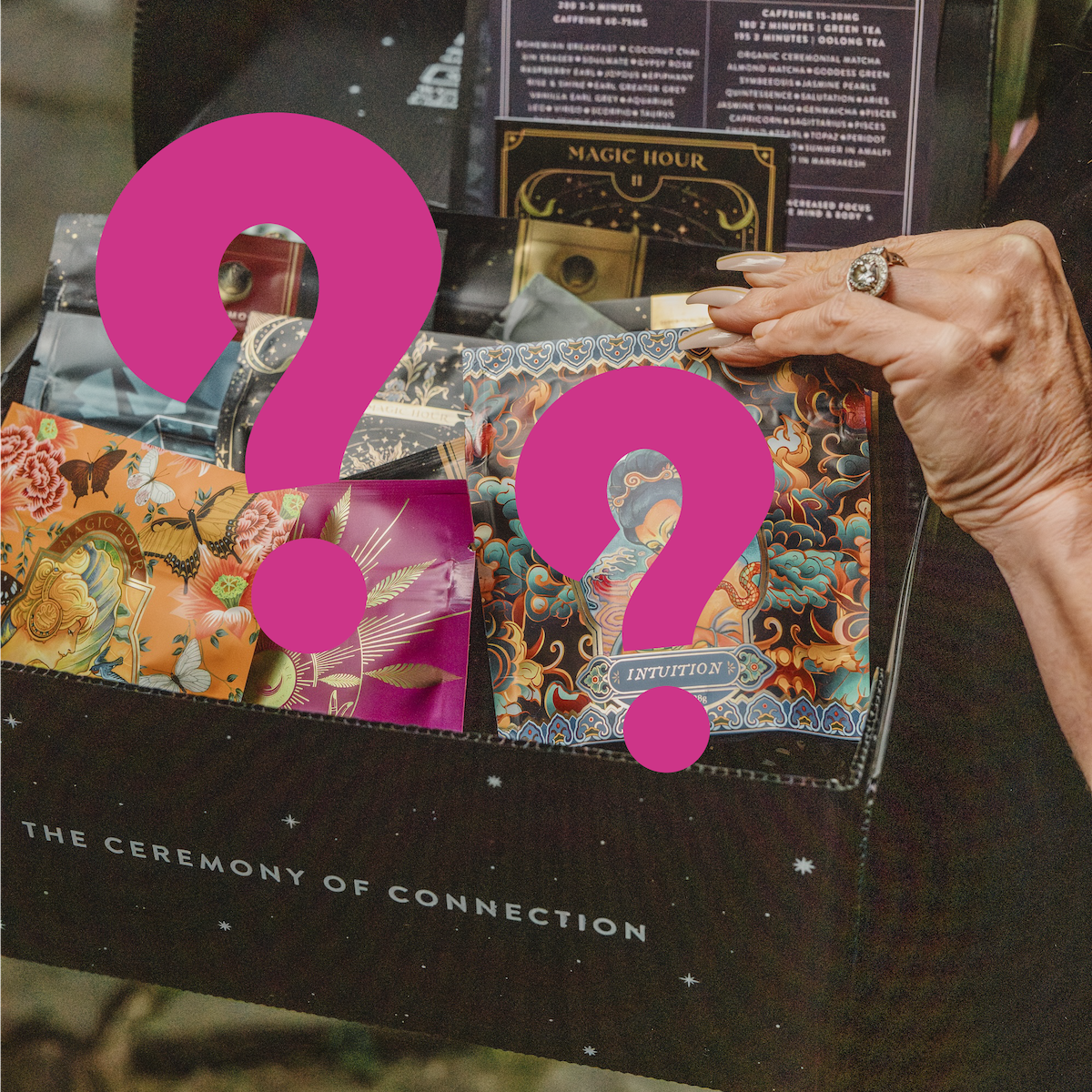 A person holds a black box labeled &quot;The Ceremony of Connection,&quot; filled with colorful tarot decks and envelopes with artistic designs. Some feature bold magenta question marks, hinting at surprises like luxurious teas or the Super Spring Mystery Box.