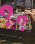 A person holds a black box labeled "The Ceremony of Connection," filled with colorful tarot decks and envelopes with artistic designs. Some feature bold magenta question marks, hinting at surprises like luxurious teas or the Super Spring Mystery Box.