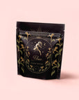 A black resealable pouch with gold and green floral designs shows a ram-headed figure and sunburst. The pale pink label reads "Aries - Garden of Eden Oolong," “Magic Hour,” and highlights Blackberry-Bergamot Earl.