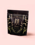 A black, resealable bag with gold and green Cancer-themed artwork featuring celestial accents and the text "Cancer: Queen of Cool Mango-Ginger-Cream White Tea" highlights mango and organic ginger against a pale pink background.