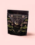 A black stand-up pouch with gold and pastel illustrations of a fish, lotus flowers, stars, and moons. The label reads "Pisces: Magnolia-Jasmine Blueberries & Cream Tea with Lion's Mane" in elegant script on a light pink background.