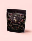 A black pouch with gold and pink centaur archer designs, labeled "Sagittarius Tea of Good Fortune & Abundance," features stars and florals and highlights its pomegranate ginger blend, set against a pale pink background.