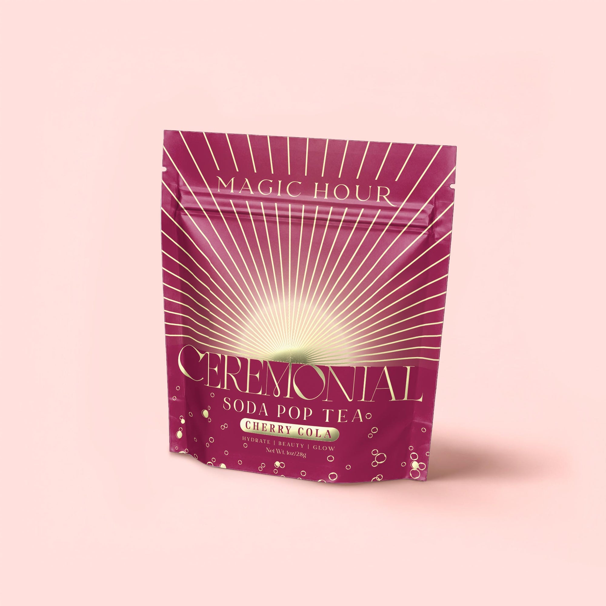 A magenta pouch labeled "Cherry Cola Black Tea: Ceremonial Soda Pop Tea" by Magic Hour features gold and white rays, bubbles, and a soft pink background—a delightful cherry cola tea with natural sweetener.