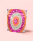 A vibrant, resealable pouch with a lively starburst design in pink, orange, and green says "333: Mango-Lemon Balm-Rose Herbal Tea with Passionflower" and stands upright on a pale pink background.