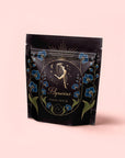 A black pouch with gold and blue floral designs features a woman pouring water, stars, and "Aquarius: Visionary Goddess Tea," hinting at the vanilla-infused black tea inside, set on a pale pink background.