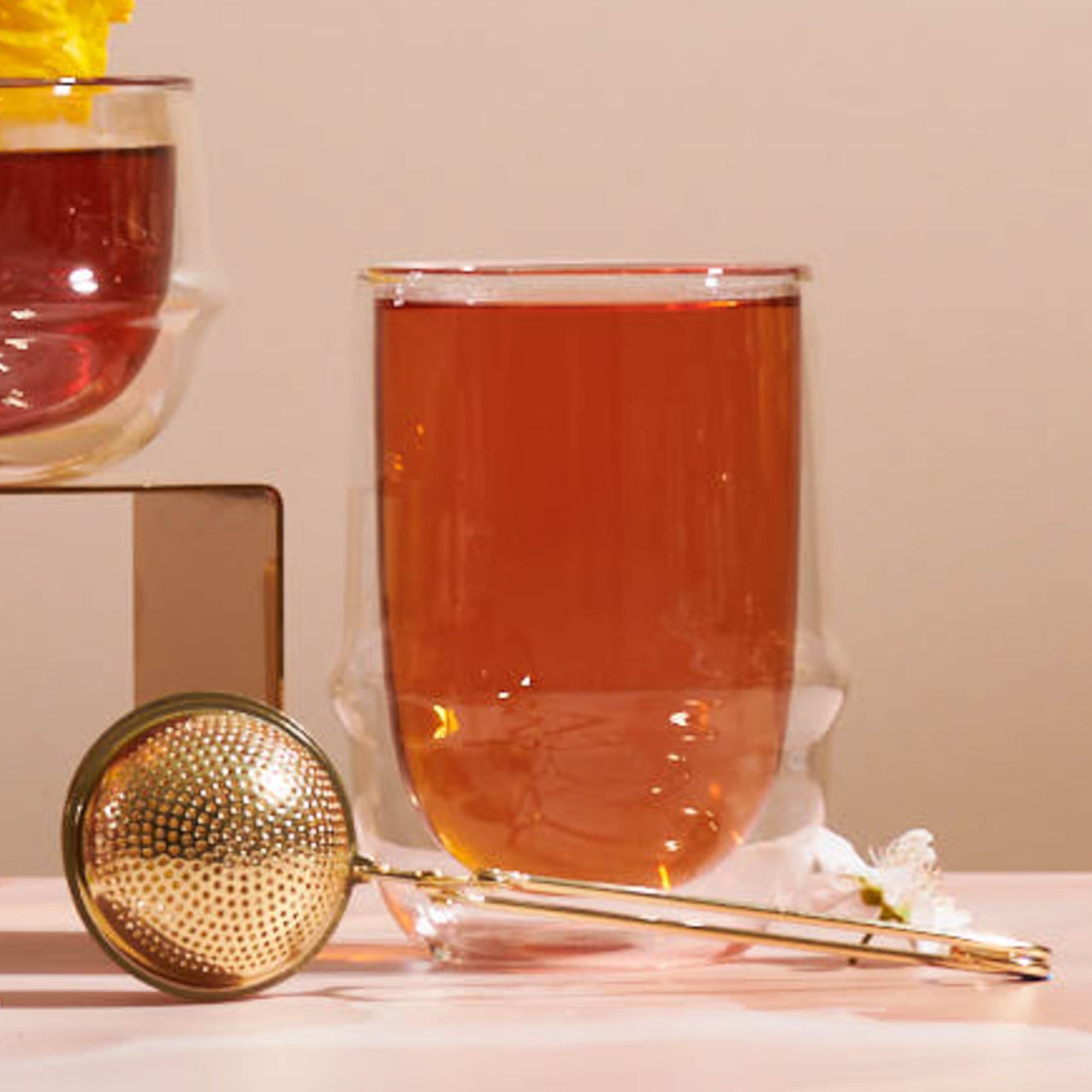 A glass of tea sits on a table beside a Luxe Loose Leaf Strainer and a small white flower, with another cup partially visible in the background.