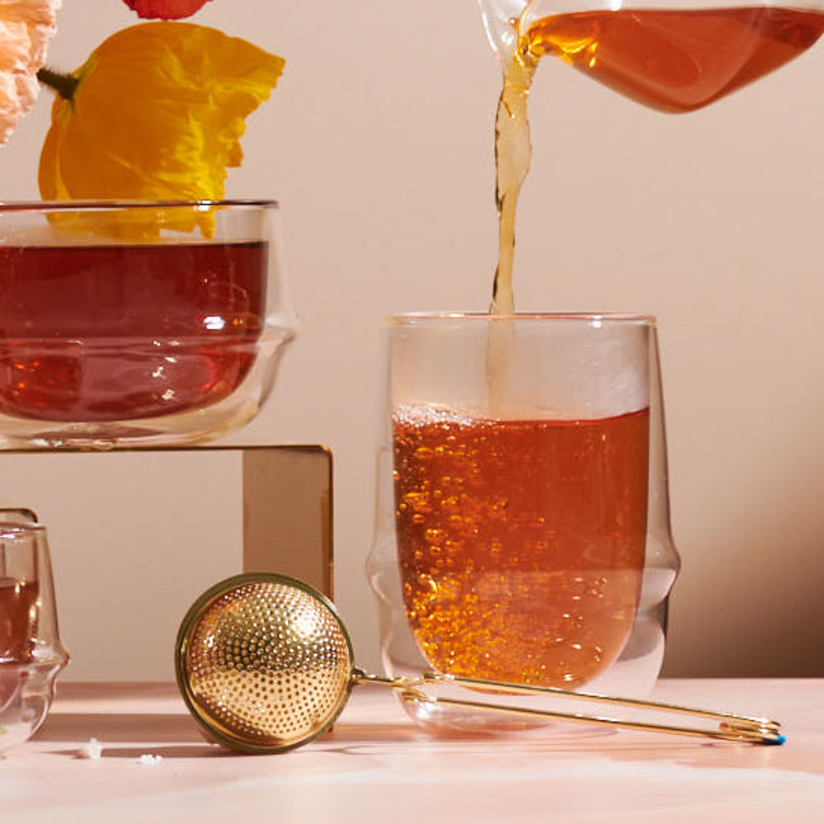 Tea is being poured into a double-walled glass using the Luxe Loose Leaf Strainer, with additional glass cups nearby. A yellow flower adorns one of the glasses, all arranged on a clear platform in soft lighting.