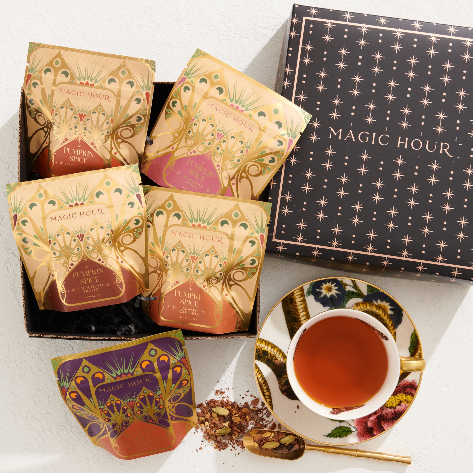 An open Best of Fall Box reveals four ornate packets of Magic Hour tea, including the delightful Pecan Pie Chai, each featuring intricate designs. Below the box, you’ll find a teacup filled with tea on a saucer, accompanied by a gold spoon and some scattered loose tea leaves. The set also includes an empty purple teacup adorned with an elaborate design.