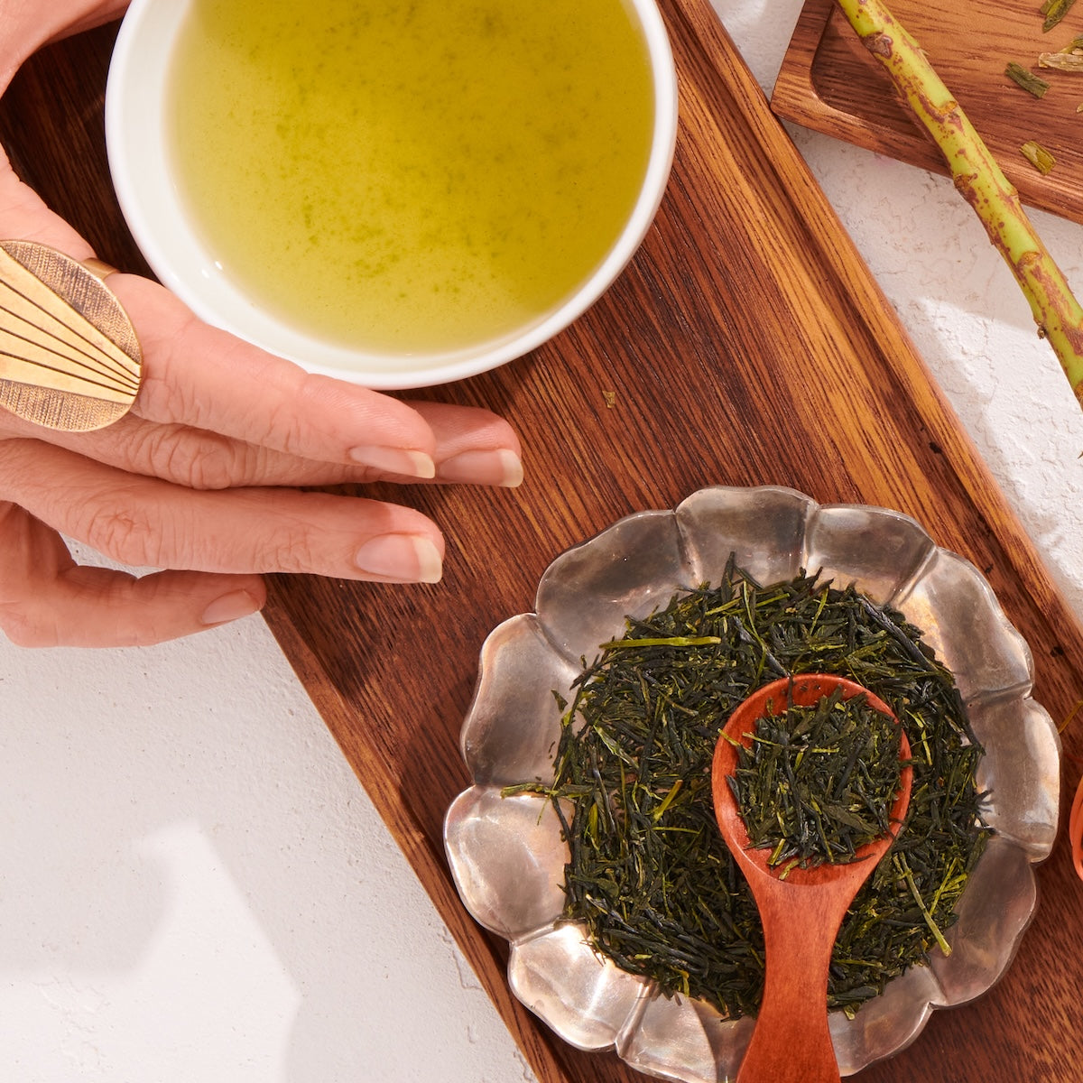 A manicured hand rests by a cup of Japanese green tea and a metal dish of Flower Fragrance Iccha Kariban Green Tea leaves on a wooden board. A wooden spoon sits atop the vibrant loose leaves, highlighting this premium Kagoshima tea.