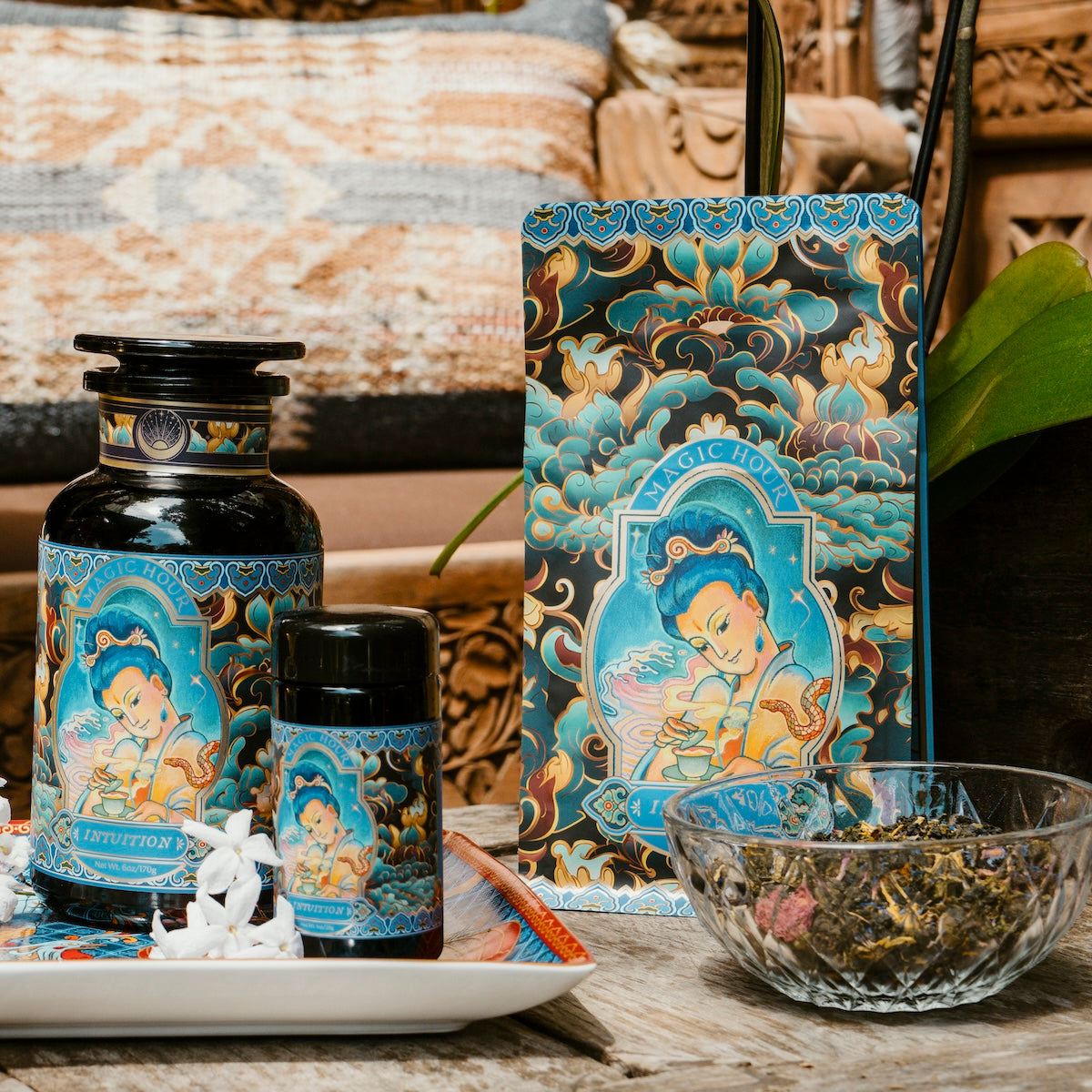 A collection of Magic Hour tea products is artfully arranged on a wooden surface, featuring the Nuwa Tea for Intuition: Dragon Yuzu Oolong Case Packs. The display includes a large jar, a smaller container of Organic Dragon Phoenix Jasmine Pearls, and a bag of tea, all adorned with intricate, colorful designs. To complete the setup, there's a bowl of loose-leaf tea and a small tray with flowers.