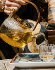 A person pours Dragon Yuzu Oolong tea from a glass teapot into a clear glass on a wooden table. The colorful tray beneath the glass adds visual interest, as the person, adorned in a flowy garment with an attention-grabbing ring and bracelet, completes the ritual. The background is filled with comfortable pillows and artistic touches, subtly evoking the calming aroma of Japanese Yuzu Essential Oil from Magic Hour’s Nuwa Tea for Intuition line.