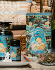 A collection of Magic Hour tea products is artfully arranged on a wooden surface, featuring the Nuwa Tea for Intuition: Dragon Yuzu Oolong Case Packs. The display includes a large jar, a smaller container of Organic Dragon Phoenix Jasmine Pearls, and a bag of tea, all adorned with intricate, colorful designs. To complete the setup, there's a bowl of loose-leaf tea and a small tray with flowers.