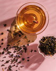 A transparent glass teacup filled with amber-colored Nuwa Tea for Intuition: Dragon Yuzu Oolong from Magic Hour sits on a pink surface. Next to it, a small ornate dish holds loose tea leaves. Some tea leaves are scattered around the dish, and the shadows create a textured pattern on the table.