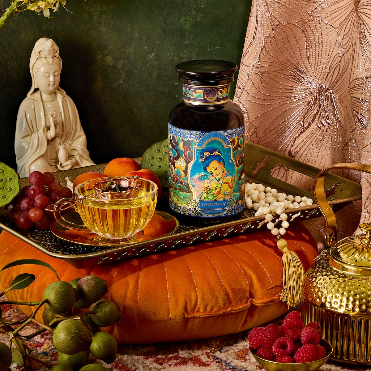 A decorative setting showcases a jar with vibrant illustrations, a clear teacup filled with Nuwa Goddess of Creation Tea for Intuition: Dragon Yuzu Oolong on a tray, surrounded by grapes, apricots, and a golden teapot. A statue and patterned cushions enhance the scene against a textured backdrop.