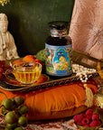 A decorative setting showcases a jar with vibrant illustrations, a clear teacup filled with Nuwa Goddess of Creation Tea for Intuition: Dragon Yuzu Oolong on a tray, surrounded by grapes, apricots, and a golden teapot. A statue and patterned cushions enhance the scene against a textured backdrop.