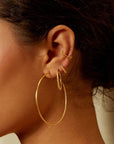 Close-up of an ear adorned with multiple gold earrings. The ear features an Ongoing Energy Large Hoop Earring by Magic Hour in the lower lobe, with two smaller hoops and a delicate ear cuff positioned on the upper ear cartilage. The hammered 18KT gold plate catches the light beautifully against the neutral background, highlighting the jewelry.